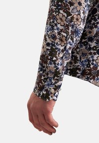 Camicia floreale a maniche lunghe con una combinazione di fiori blu, marroni e crema su uno sfondo chiaro; presenta bottoni ai polsini.