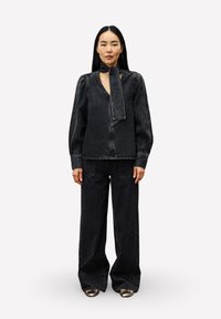 Zwart denim outfit met een blouse met lange mouwen en een halsdoek, en wijde broekspijpen die een ontspannen pasvorm en subtiele stikseldetails tonen.