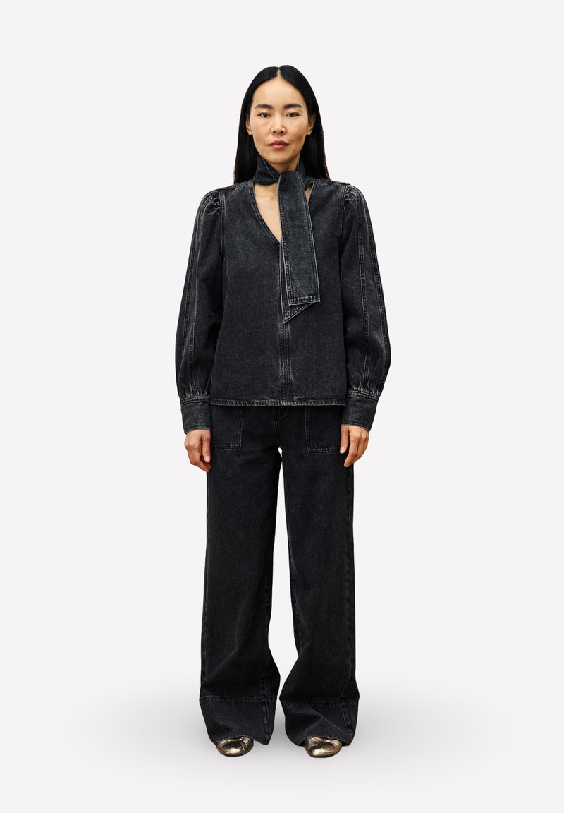 Zwart denim outfit met een blouse met lange mouwen en een halsdoek, en wijde broekspijpen die een ontspannen pasvorm en subtiele stikseldetails tonen.