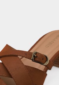 Καφέ δερμάτινο σανδάλι με σταυρωτά λουριά και χρυσή αγκράφα σε τακούνι πλατφόρμα.