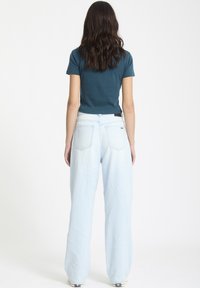Lysblå vide jeans og en tætsiddende teal kortærmet skjorte. Jeansene har et klassisk fem-lomme design og synlige syninger.