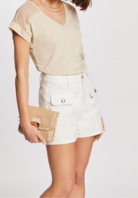 Top beige de cuello en v con mangas transparentes y estampadas, combinado con pantalones cortos blancos con bolsillos delanteros. Sosteniendo un bolso de mano tejido de paja.