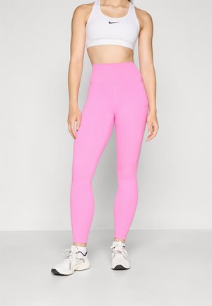 Leggings roses taille haute en matériau extensible, avec une texture lisse et des poches latérales, associés à un brassière de sport blanche et des baskets.