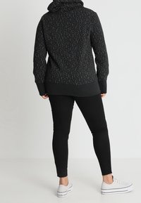 Persona che indossa un hoodie nero a motivi, pantaloni skinny neri e sneakers bianche basse, in piedi contro uno sfondo semplice, mostrata di spalle.