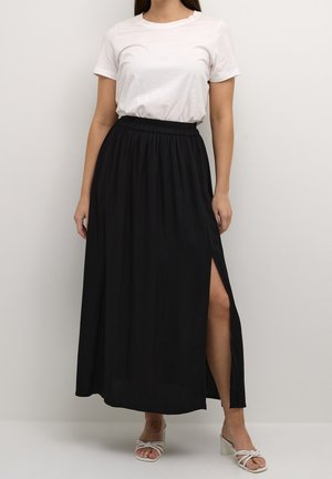 Black maxi skirt with a side slit, elastic waistband, flowing fabric, paired with a white t-shirt and light-colored heeled sandals.