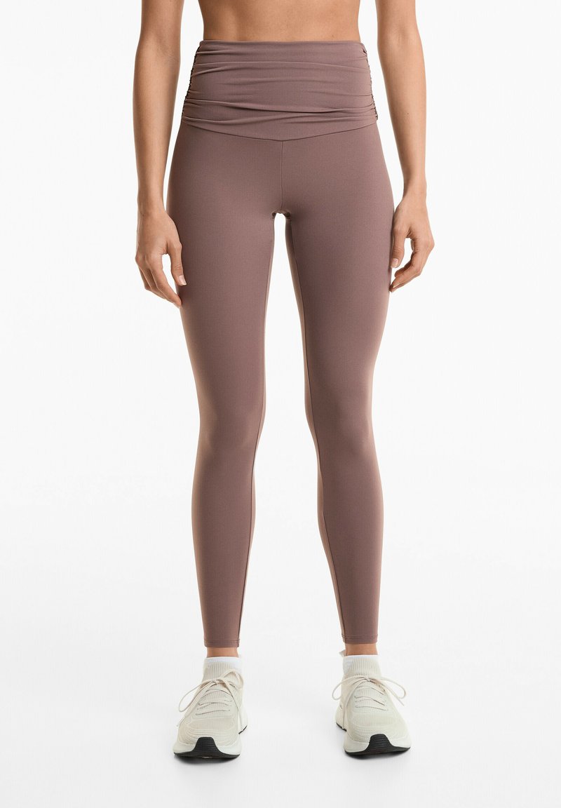 Leggings cu talie înaltă, ajustate, dintr-un material moale de culoare mauve, cu detalii plisate la talie, asortate cu pantofi sport de culoare deschisă.