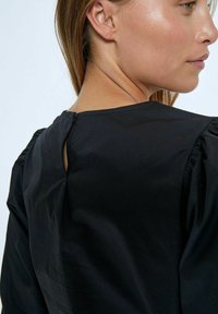 Blusa nera con maniche a sbuffo e chiusura a oblò sulla schiena. Tessuto morbido con una leggera lucentezza, design minimalista.