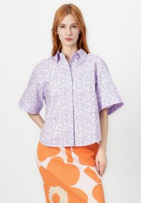 Chemise à manches courtes en tissu violet clair avec un motif floral en contour. Le modèle la porte avec une jupe à motifs orange.