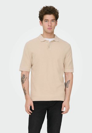 Mladý muž s kučeravými vlasmi, oblečený v béžovej krátkemu rukávu polo tričku a čiernych nohaviciach, ukazuje tetovania na oboch predlaktiach, stojaci pred jednoduchým pozadím.