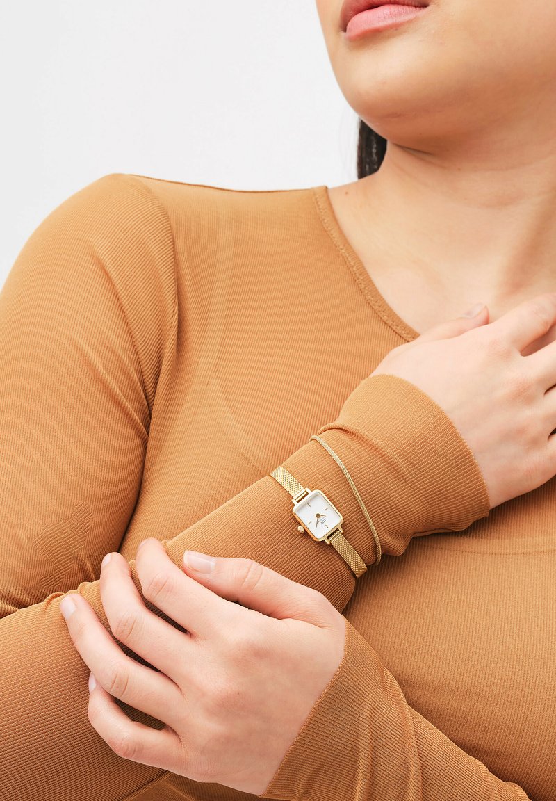 Goldfarbene Mesh-Armbanduhr mit einem quadratischen weißen Ziffernblatt, getragen an einem Handgelenk. Der Träger trägt ein braunes, langärmliges Oberteil mit Rippstruktur und einem einfachen Design.