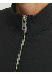 Sort zip-jakke med høj krave, ribbet tekstur og metalzipperdetaljer. Har en glat stofoverflade.
