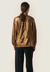 Blusa di bronzo metallico con colletto alto e maniche lunghe, caratterizzata da un design drappeggiato sul retro e una texture liscia. Abbinata a pantaloni neri.