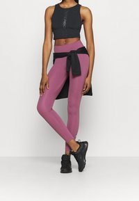 Czarny sportowy crop top z logo, w połączeniu z wysokim stanem śliwkowymi legginsami. Na sobie ma czarną związana kurtkę i czarne buty sportowe.