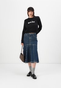 Felpa nera corta con testo grafico bianco, abbinata a una gonna di denim di lunghezza media con orlo sfrangiato e due tasche laterali. Mocassini neri e calzini grigi.