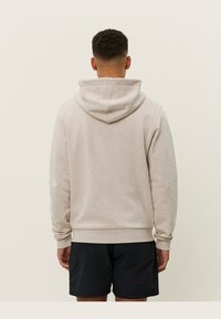 Sudadera color crema con capucha ajustable, puños y dobladillo acanalados. La tela parece suave con una textura lisa. Llevada sobre pantalones cortos oscuros.