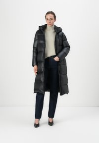 Zwarte pufferjas met capuchon, voorzien van een glanzende textuur en een doorgestikte ontwerpen, gecombineerd met een lichte trui en donkere jeans.