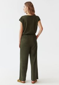 Salopetă de culoare verde măslinie cu mâneci scurte, croială lejeră, talie elastică și două buzunare pe spate; fabricată dintr-un material moale și neted.