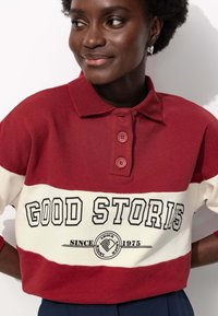 Polo rayé rouge et crème avec un col, trois boutons et un design graphique avec le texte "BONNES HISTOIRES DEPUIS 1975."