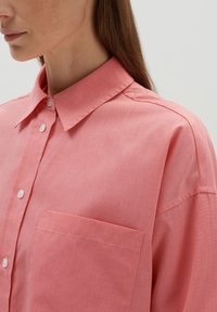 Chemise boutonnée rose clair avec un col, dotée d'une poche poitrine et d'un tissu texturé. Gros plan mettant en avant les détails de couture.