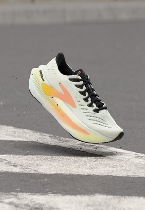 Witte Brooks hardloopschoen met zwarte veters en oranje-gele verlooplogo zwevend boven een asfaltweg met witte strepen.