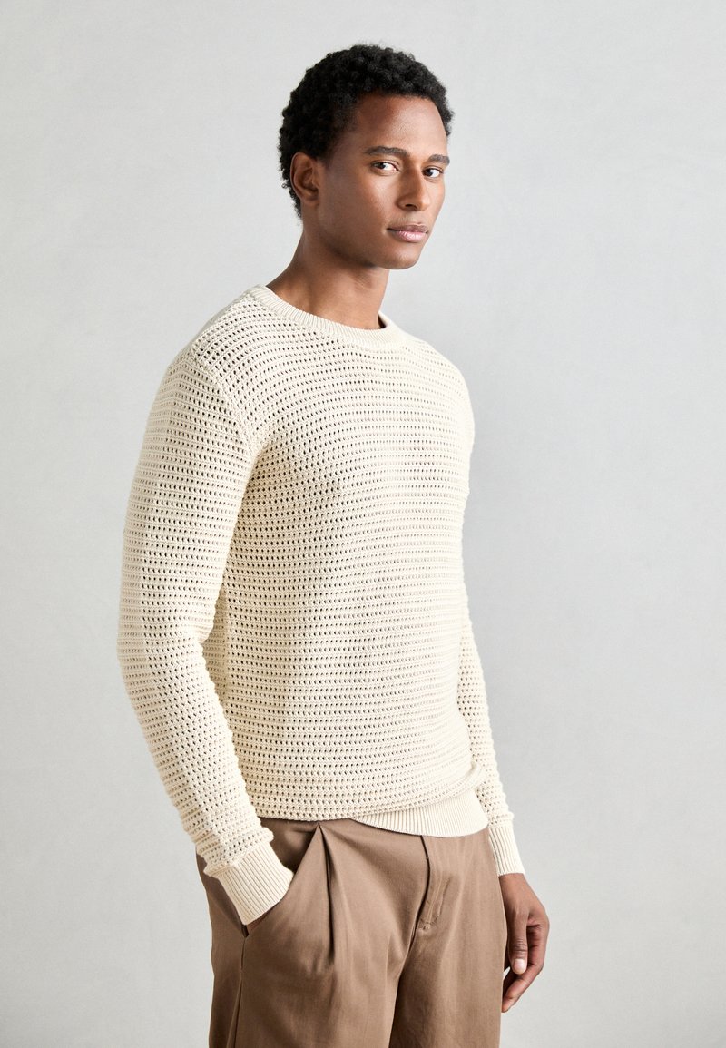 Pull en tricot crème avec un design texturé à mailles ouvertes et des manches longues, associé à un pantalon marron clair.