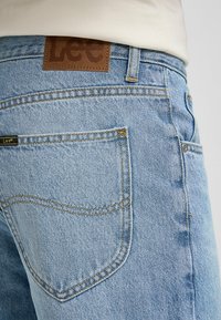 Ljusblå denimjeans har en brun läderning med texten "Lee", med synlig sömnad och en klassisk femficksdesign.