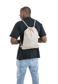 Batoh na stažení vyrobený z jemného béžového materiálu s měkkou texturou. Obsahuje nášivku s logem a nastavitelné popruhy, nošený na černém tričku.