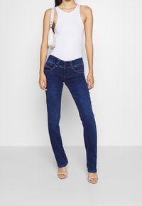 Modré džínové slim-fit kalhoty s pěti kapsami a světlým praním. Kombinovány s bílým tílkem a malou bílou kabelkou přes rameno.