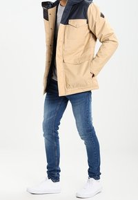 Chaqueta beige y azul marino con capucha, que cuenta con bolsillos frontales y cierre de botones; combinada con jeans ajustados y zapatillas blancas.