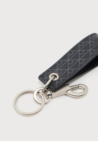 Μαύρο δερμάτινο λουράκι με σχέδιο, προσαρτημένο σε ασημένιο κρίκο κλειδιών και κούμπωμα αστακού σε λευκό φόντο.