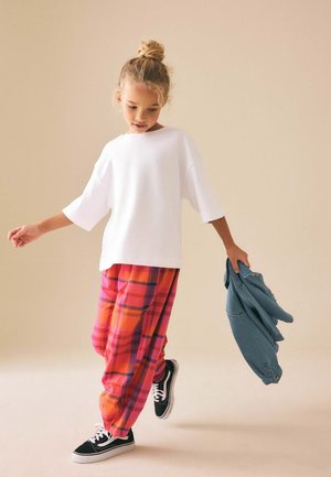 Young girl wearing white oversized shirt, bright pink and orange plaid pants, holding a blue denim jacket, standing on one foot.
