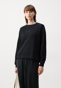 Sudadera negra fabricada en tejido suave, con cuello redondo, puños acanalados y un logo simple en el pecho. Combinada con pantalones a juego.