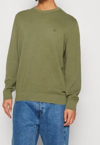 Osoba ubrana w oliwkowozielony sweter z długim rękawem ze ściągaczami na mankietach i dole, w połączeniu z niebieskimi dżinsami o luźnym kroju na jednolitym tle.