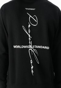 Svart hettegenser med hvit tekst "Project Care" og "WORLDWIDE STANDARD" med et kryssdesign. Myk bomullsblanding med ribbestrikkede mansjetter.