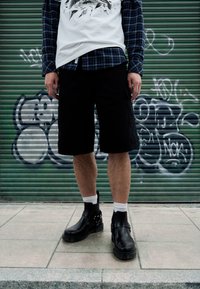 Pantaloni scurți negri cargo cu buzunare laterale, tricou alb cu imprimeu, cămașă albastru în carouri și ghete negre de piele până la gleznă cu curele, purtate peste șosete albe.