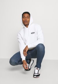 Sudadera blanca con capucha y un logo negro de Levi's, combinada con unos vaqueros ajustados azules y zapatillas negras con acentos blancos y rojos.