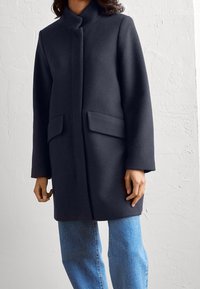 Mörk ullkappa med hög krage, framknäppning och två sidofickor. Kombination med ljusblå jeans med en slät yta.