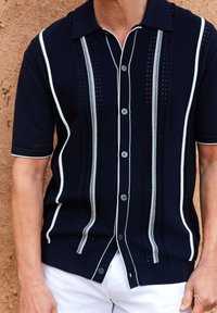Camisa de manga corta navy con rayas verticales blancas, detalles texturizados intrincados y cierres de botón. Cuello y puños acanalados.