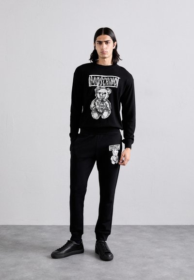 Sudadera negra de Moschino con estampado gráfico de oso de peluche y pantalones de chándal negros a juego con logotipo y gráfico del oso en el bolsillo.