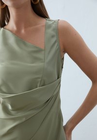 Robe sans manches de couleur vert olive en tissu lisse, présentant un décolleté asymétrique et des détails drapés à la taille. Design minimaliste.