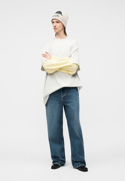 Γυναίκα που φοράει μπεζ καπέλο με μαύρο σχέδιο, στρωμένα υπερμεγέθη λευκά και απαλό κίτρινο τοπ, φαρδιά μπλε τζιν και μαύρα παπούτσια, στέκεται με σταυρωμένα τα χέρια.