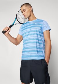 Lyseblå treningstopp med horisontale striper og korte ermer, kombinert med svarte shorts. Modellen holder en tenniskølle.