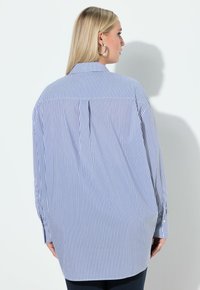 Niebiesko-biała koszula w paski na guziki, z kołnierzykiem, długimi rękawami i luźnym krojem. Z tyłu posiada plisę zapewniającą większą swobodę ruchów.