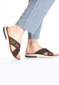Brown sandals with cross-straps, featuring a textured upper and cushioned sole. The sole has a white midsection and black tread for traction.