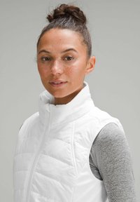 Gilet imbottito bianco con colletto alto, caratterizzato da una zip frontale e senza maniche, indossato sopra una maglietta grigia a maniche lunghe. Texture liscia.