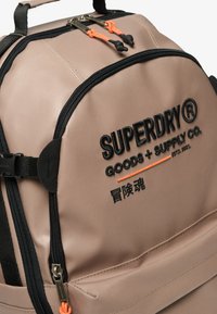 Beige ryggsäck med svarta accenter, som har en dragkedjeficka, orange dragkedjehandtag och en broderad logotyp på framsidan. Hållbart material.