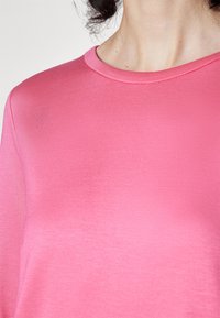 Femme portant un t-shirt rose uni à col rond, montrant le décolleté, l'épaule et le haut de la poitrine avec des cheveux foncés visibles.