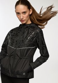 Czarna lekka kurtka z połyskującą wstawką, regulowanym kapturem, sznurkiem w talii i odblaskowymi akcentami. Posiada logo na dolnej lewej stronie.
