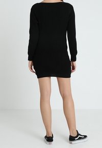 Robe noire en tricot à manches longues, avec un décolleté large et une texture côtelée, tombant au-dessus des genoux. Associée à des baskets noires.