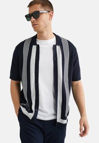 Navy knit short-sleeve shirt with gray vertical stripes, button front, and a relaxed fit, paired with a white T-shirt and sunglasses.